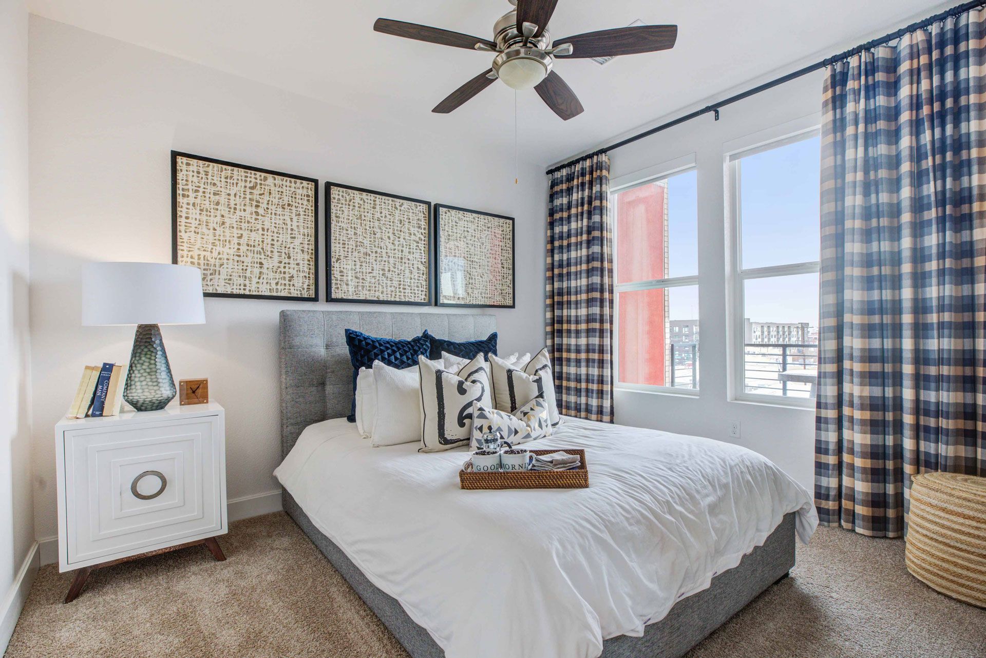Bedroom in a modern apartment with a gray upholstered bed, white bedding, blue pillows, and plaid curtains.