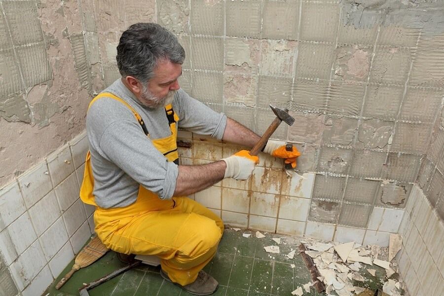 A man is kneeling down in a bathroom holding a hammer.