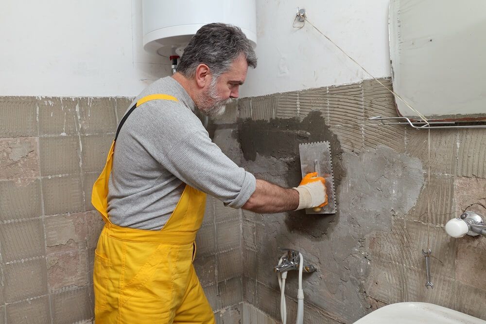 A man in yellow overalls is plastering a wall in a bathroom.