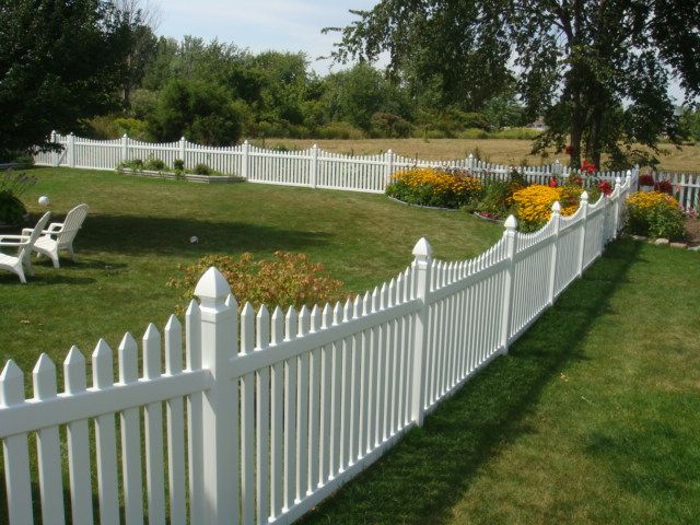 A white picket fence curves through a backyard garden filled with yellow flowers and a small patch of red blooms.