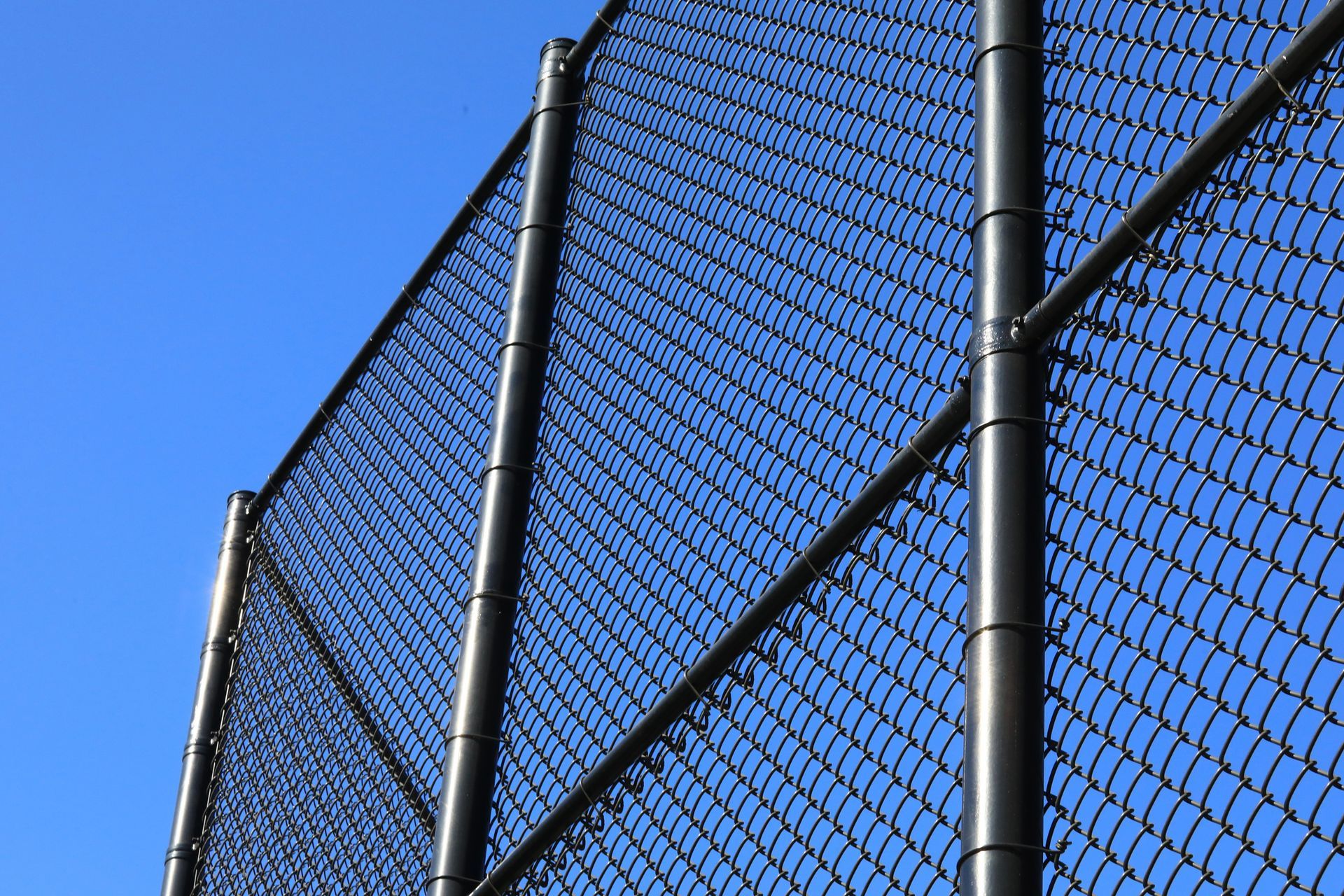 A tall chain-link fence set against a clear blue sky, with sunlight casting shadows on metal links. 