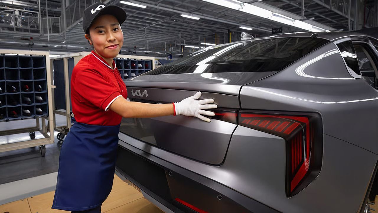 Un obrero de fábrica, vestido con una camisa roja y un guante blanco, inspecciona el maletero de un coche Kia gris mate en la línea de montaje.