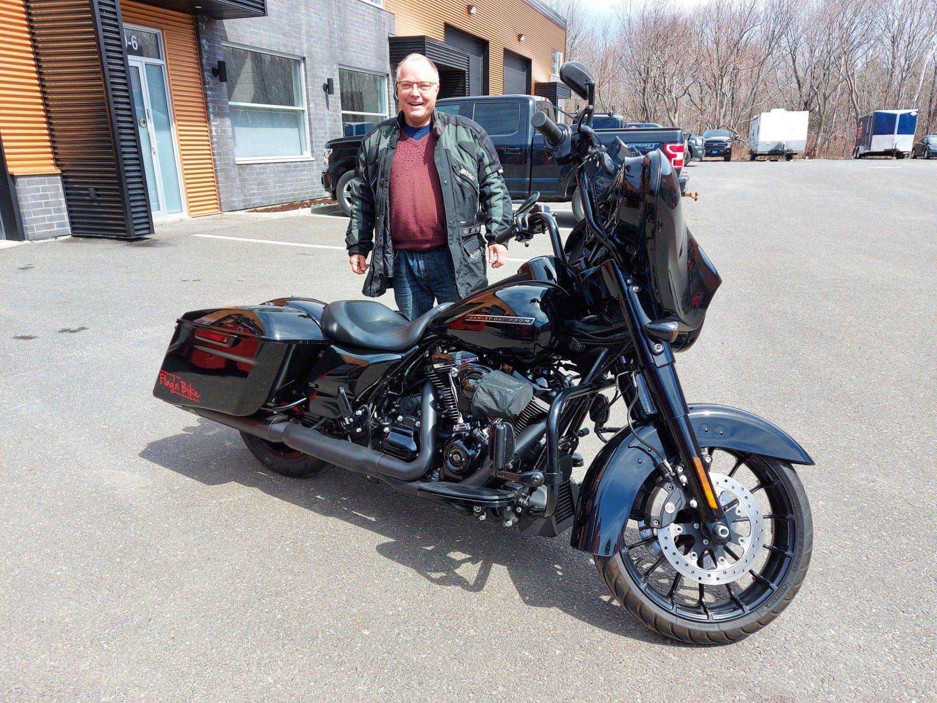 Un homme en veste de cuir pose avec une moto Harley-Davidson noire sur un parking.