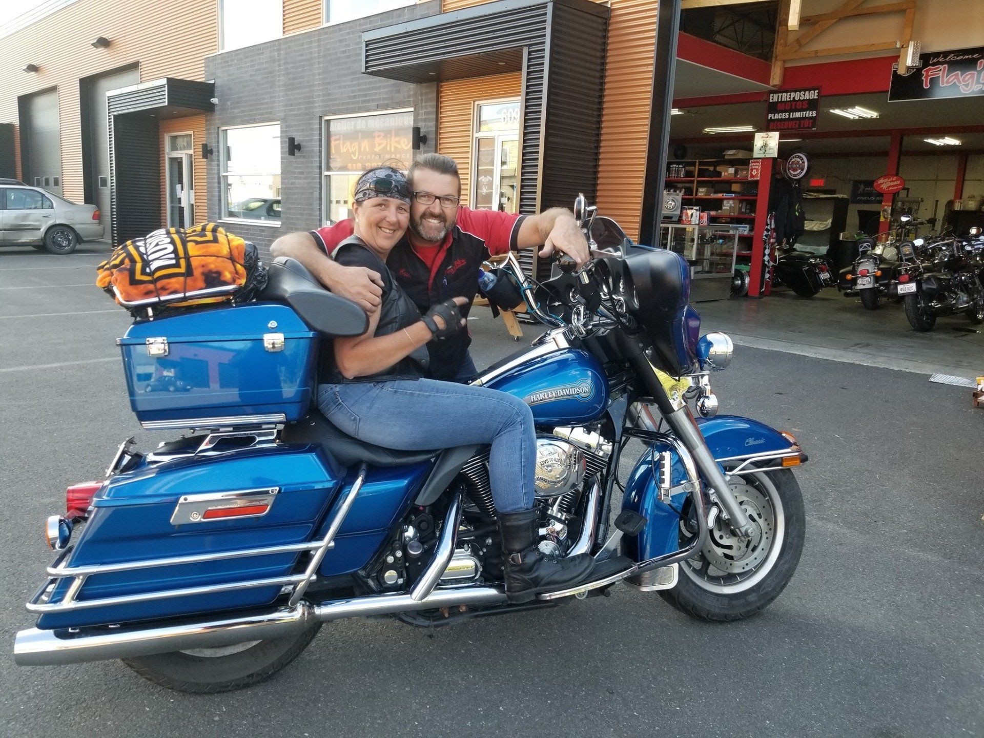 Couple sur une moto Harley-Davidson bleue, posant, souriant, devant un concessionnaire.