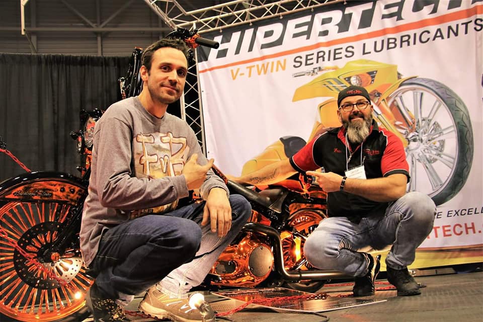 Deux hommes sont accroupis près d'une moto orange devant un stand de lubrifiants Hipertech, pointant du doigt et levant le pouce.