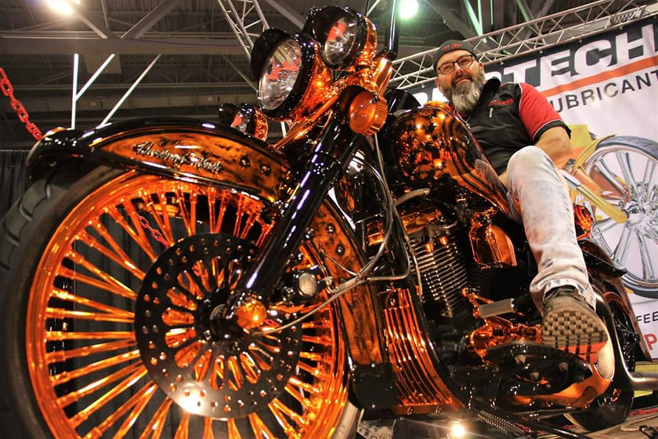 Homme assis sur une moto noire et cuivrée à grandes roues ; à l'intérieur d'un spectacle.
