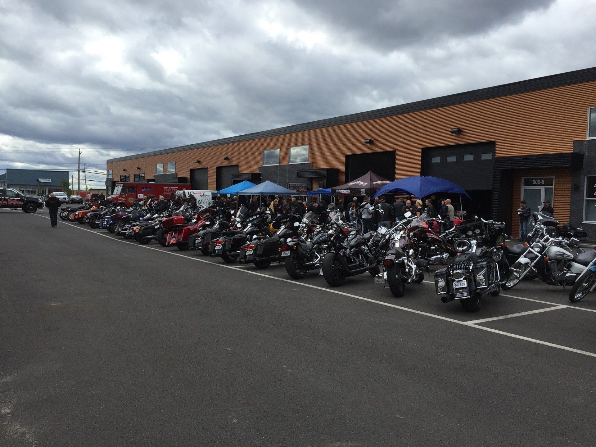Rangée de motos garées devant un bâtiment, des gens rassemblés à proximité sous des tentes par une journée nuageuse.