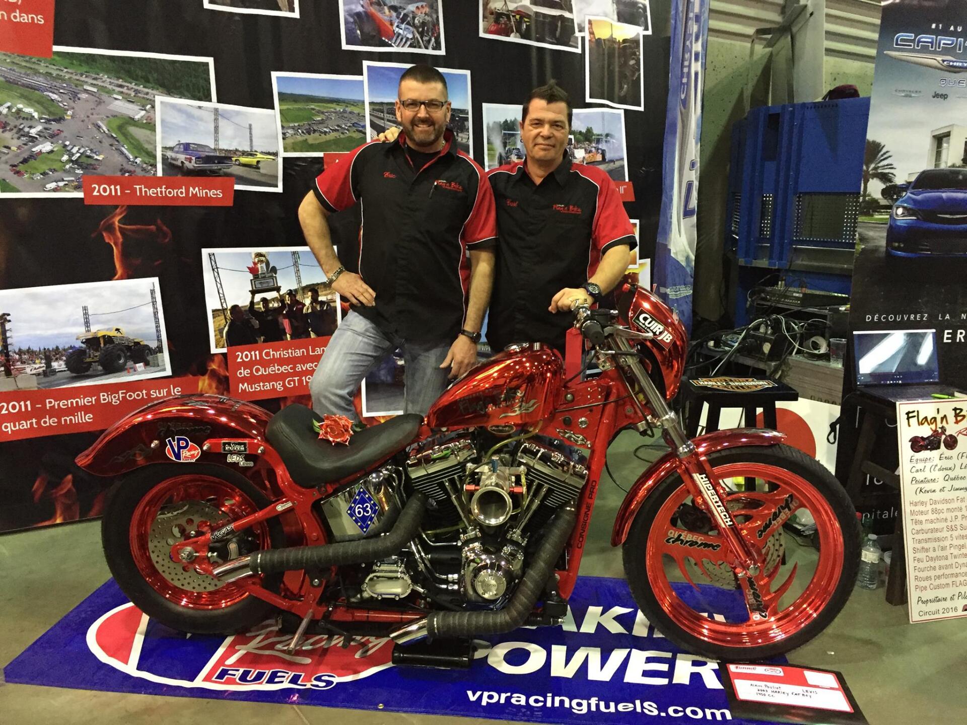 Deux hommes se tiennent sur un stand de salon avec une moto rouge customisée. Le logo « VP Racing Fuels » est visible.