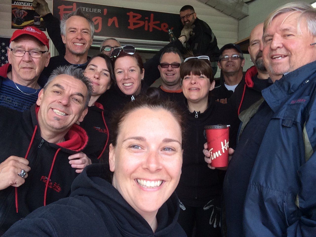 Selfie de groupe avec des personnes souriantes devant un panneau « Fly'n Bike », certaines portant des sweats à capuche rouges et tenant un café.
