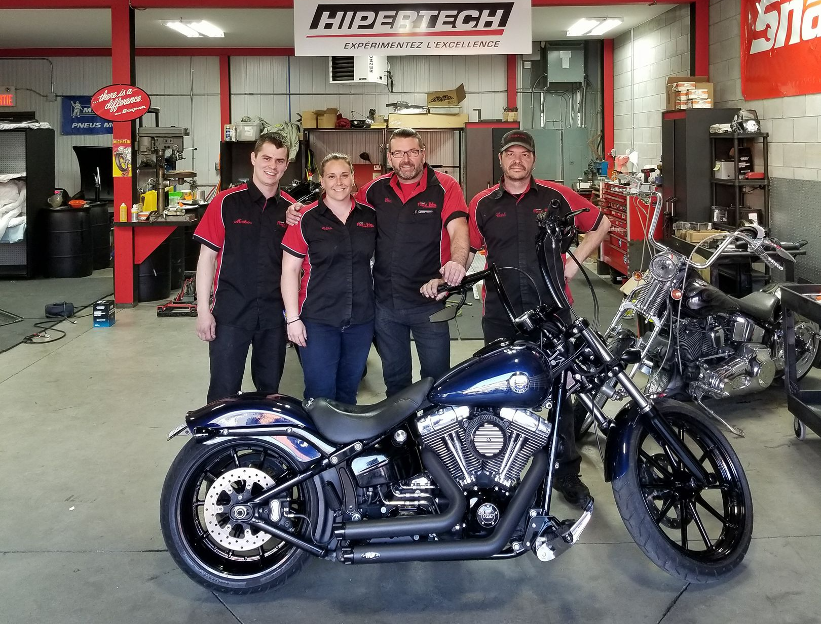 Quatre personnes posent avec une moto bleu foncé dans un atelier de réparation. L'enseigne est « HIPE TECH ».