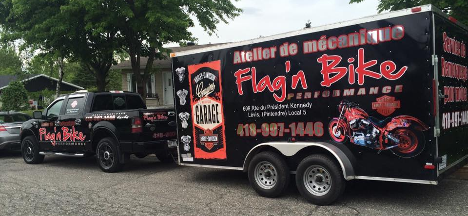 Camion noir tirant une remorque noire avec un texte rouge et blanc : garage « Flag'n Bike Performance ».