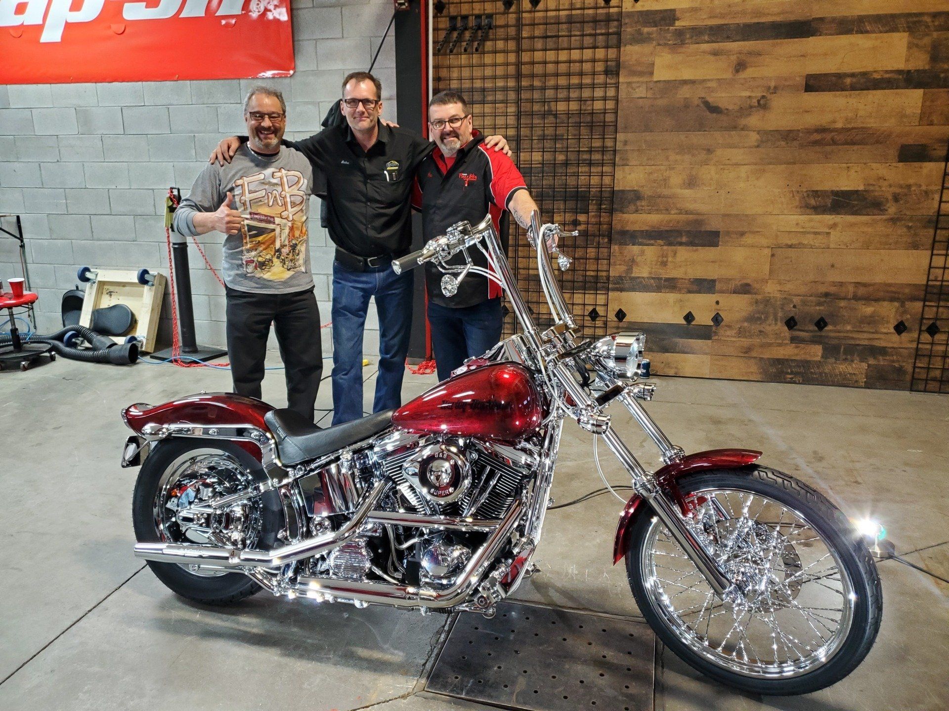 Trois hommes posent avec une moto customisée, rouge et chromée, dans un atelier.