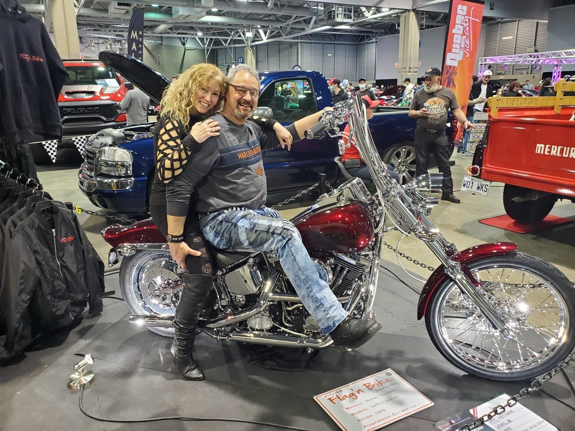 Un homme et une femme posant sur une moto personnalisée lors d'un salon automobile en salle ; un homme avec des lunettes.