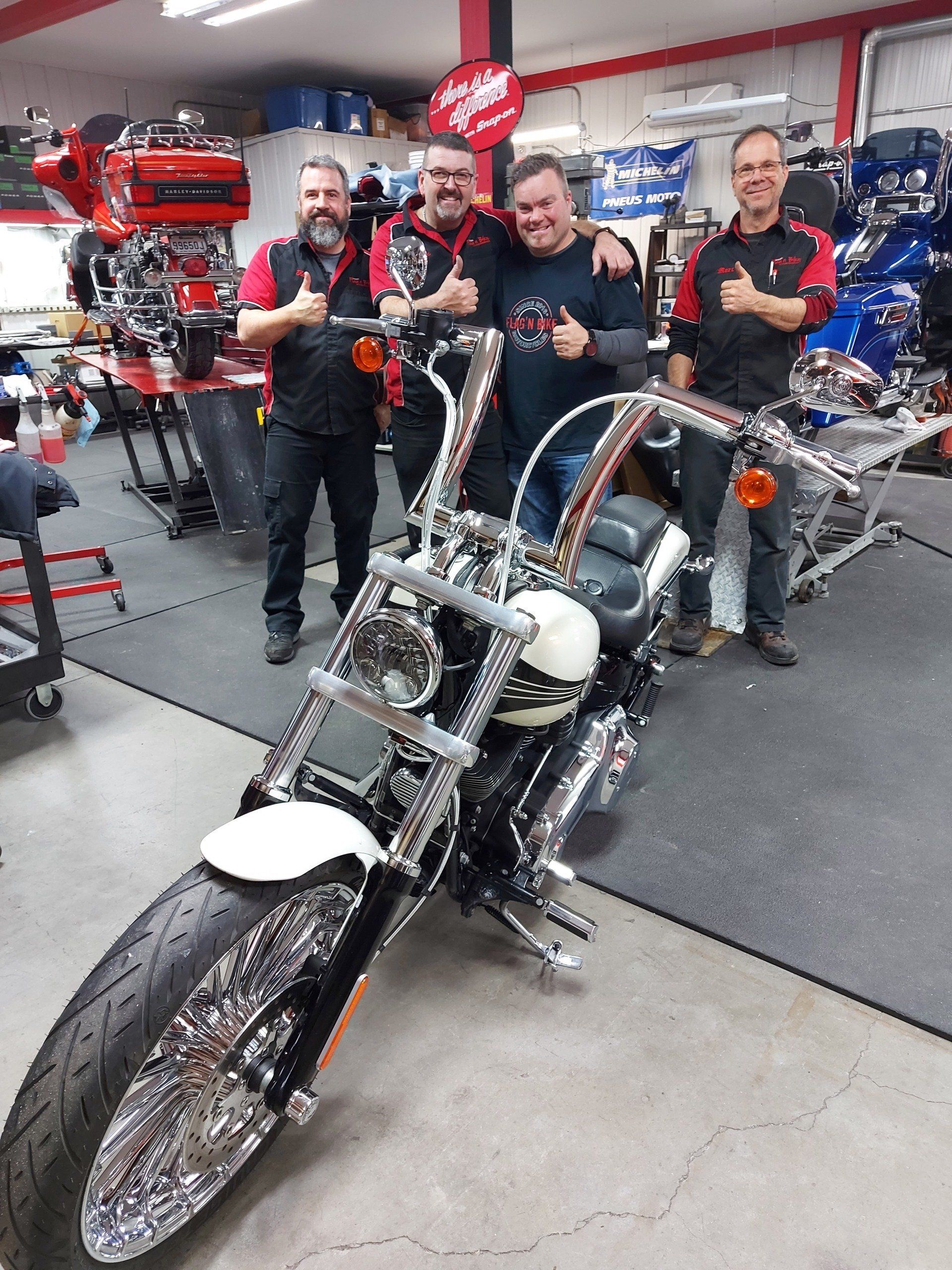 Quatre hommes posent avec une Harley-Davidson chromée dans un garage. Ils lèvent le pouce en souriant.