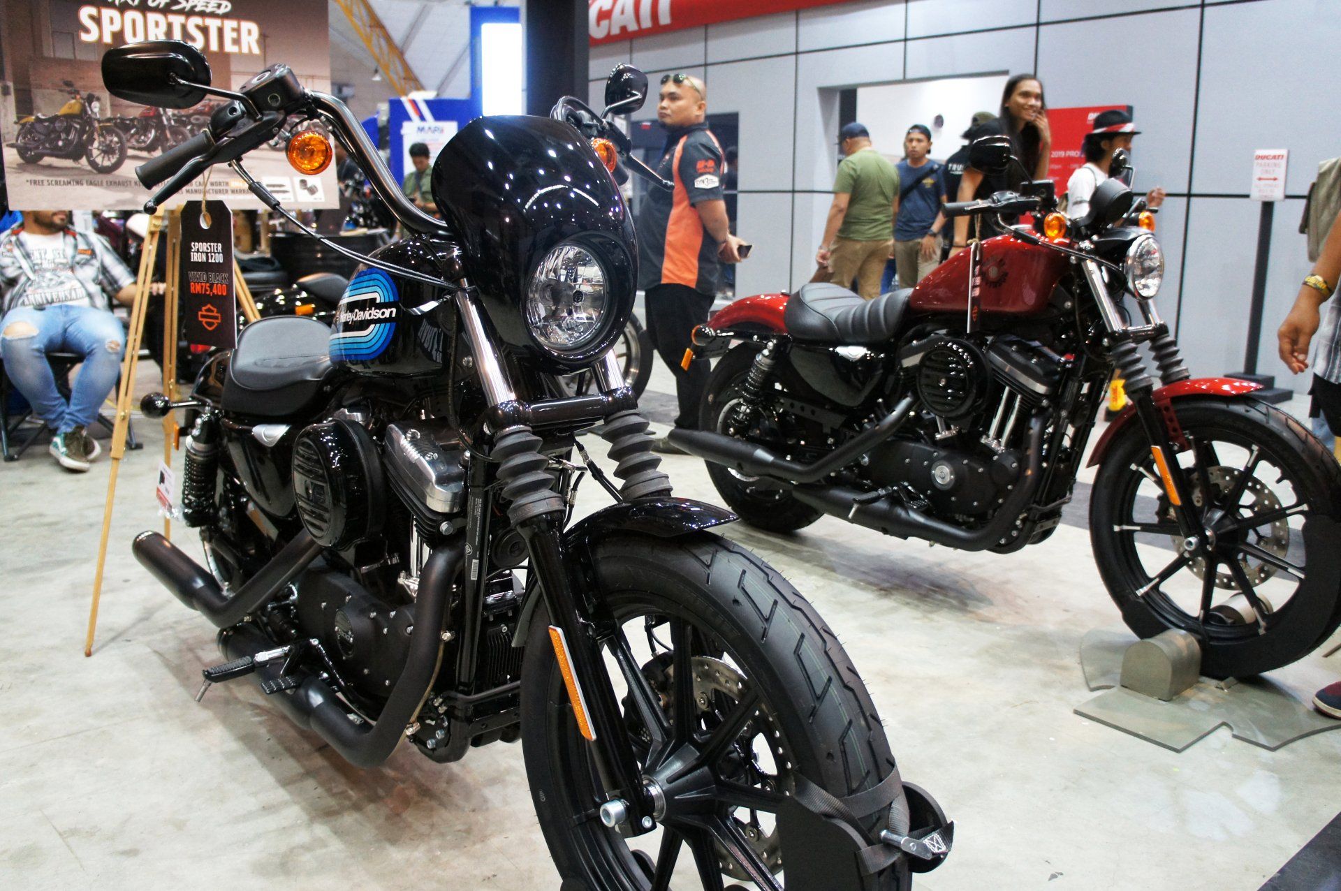 Deux Harley-Davidson exposées lors d'un événement en intérieur ; l'une noire, l'autre rouge. Des personnes sont visibles à l'arrière-plan.