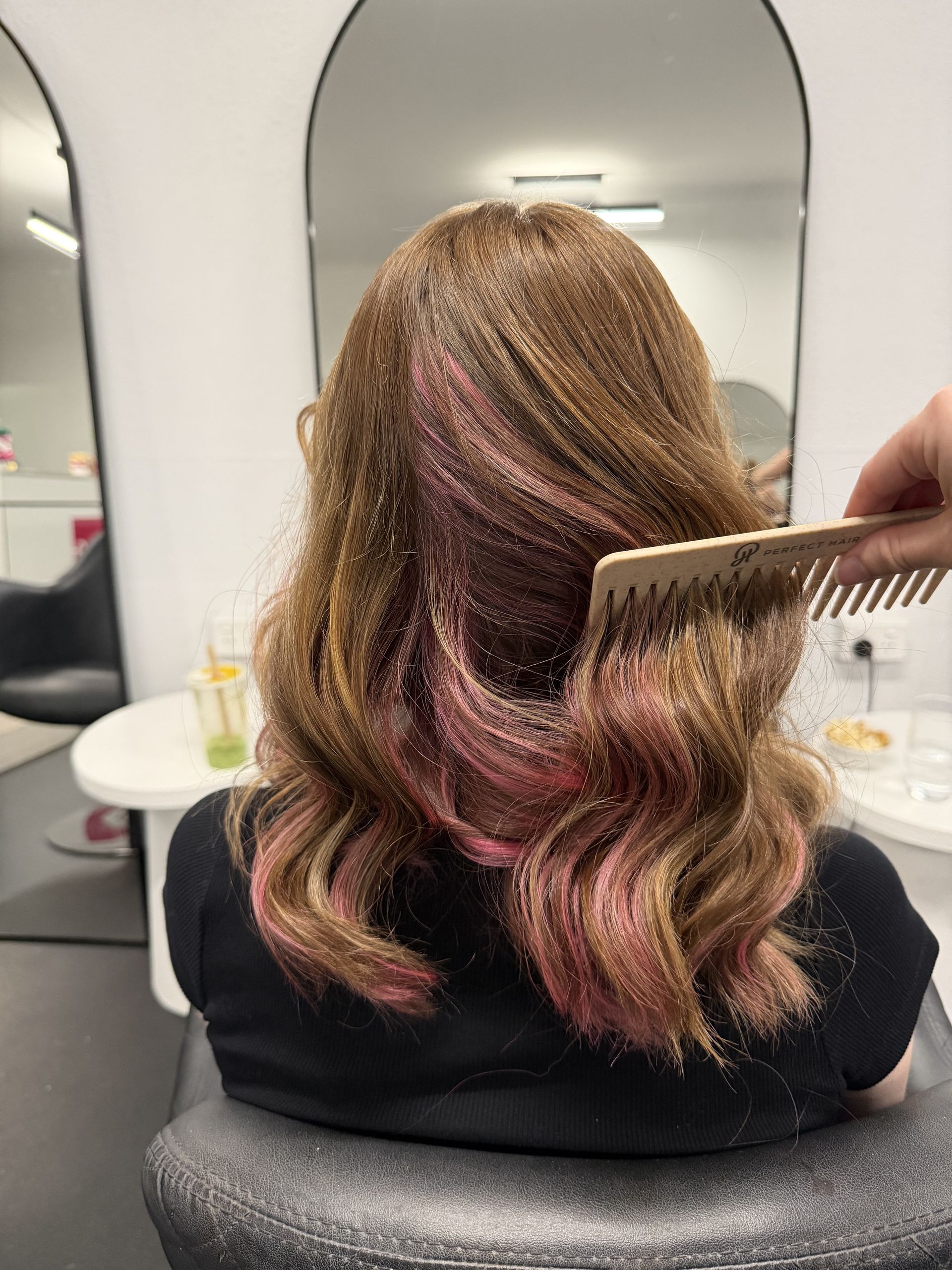 Back view of wavy shoulder-length hair with pink highlights being combed in a salon mirror  — Elysian Salon In Wyong, NSW