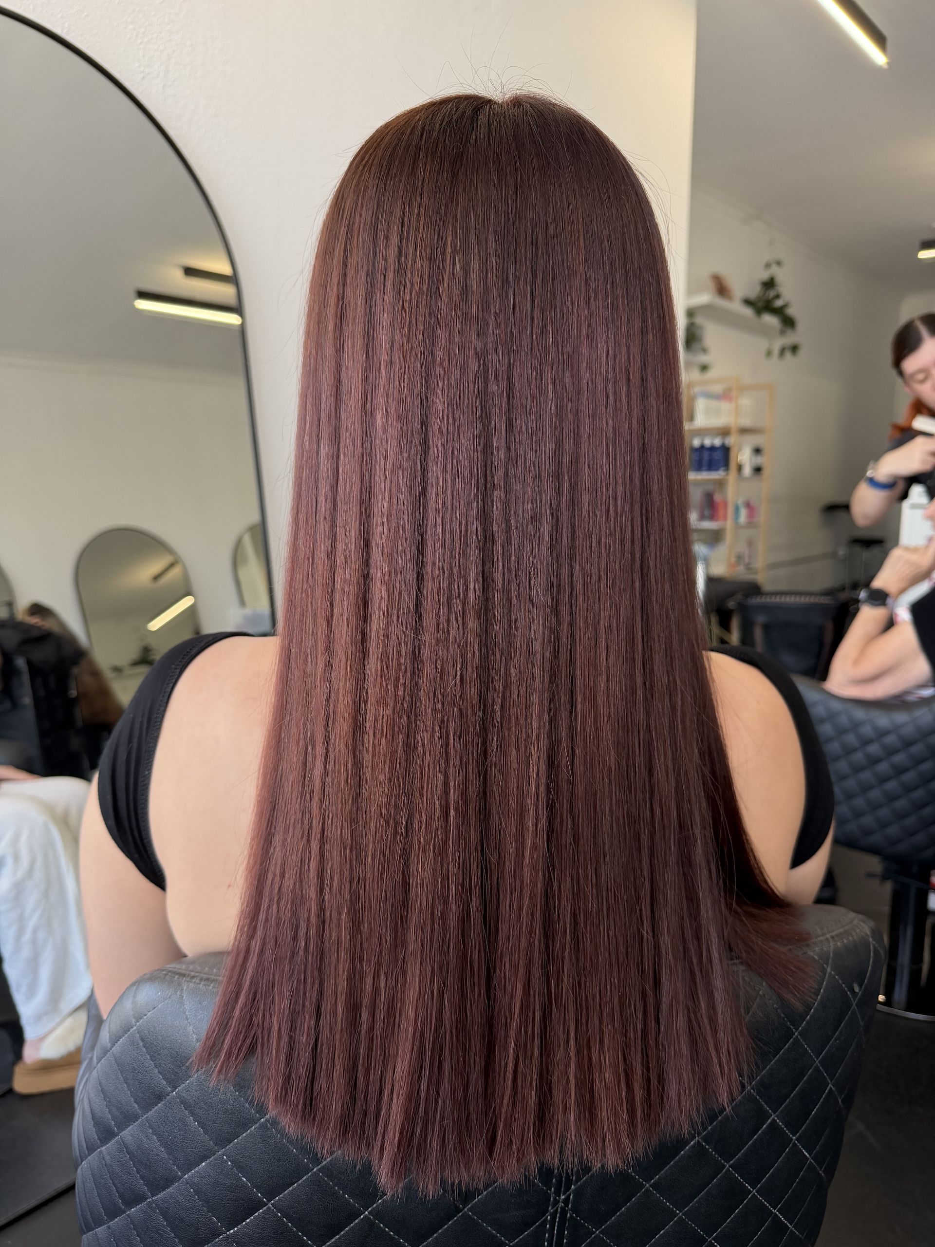 Hairdresser Applying Dye to Brown Hair With Gloved Hands and a Brush — Elysian Salon In Wyong, NSWv Straight dark brown hair down the back in a salon chair, with a mirror and stylist in the background — Elysian Salon In Wyong, NSW