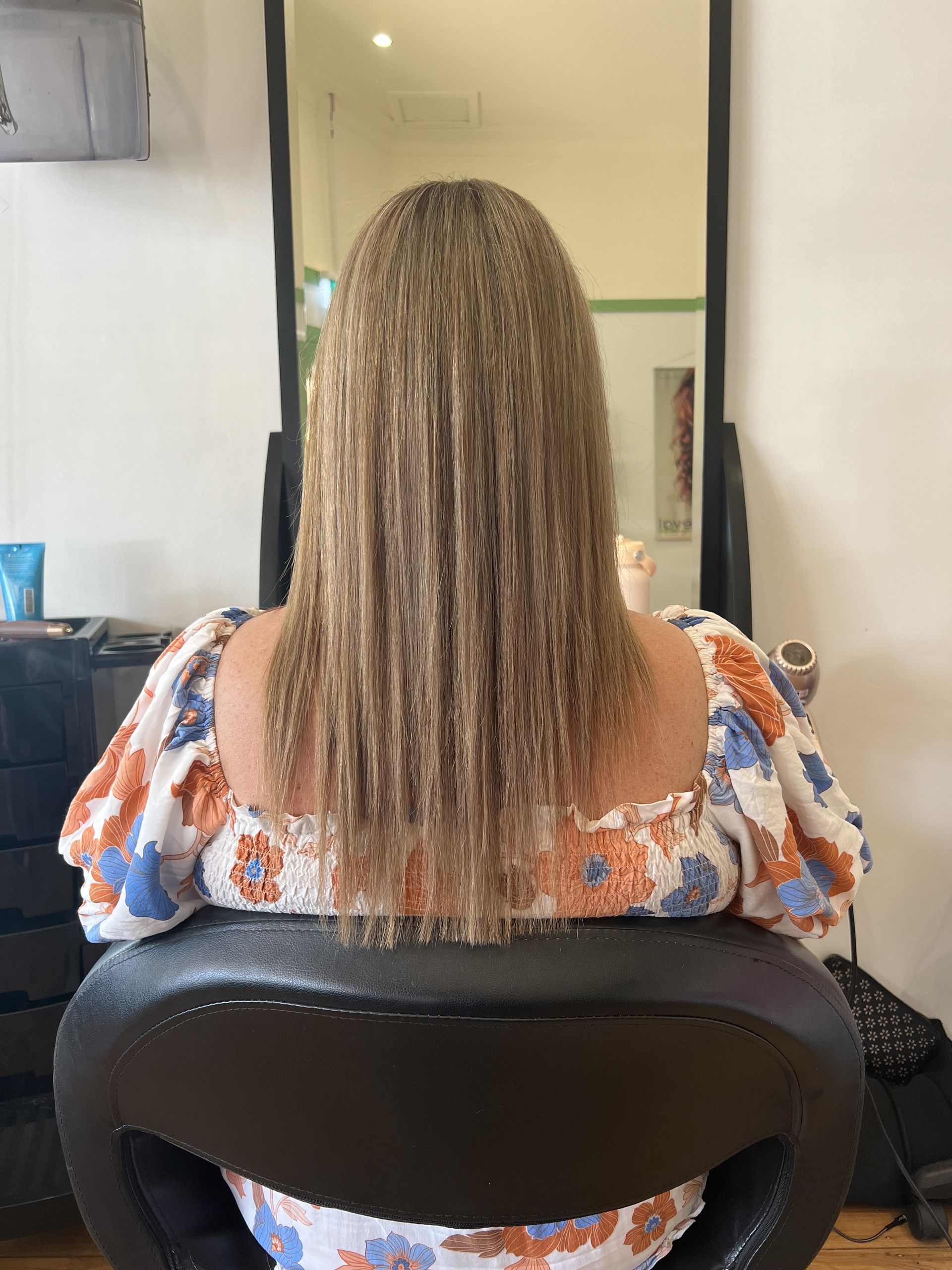 Person seated in a chair, seen from behind, with long curly hair and a floral top in a salon-like setting — Elysian Salon In Wyong, NSW