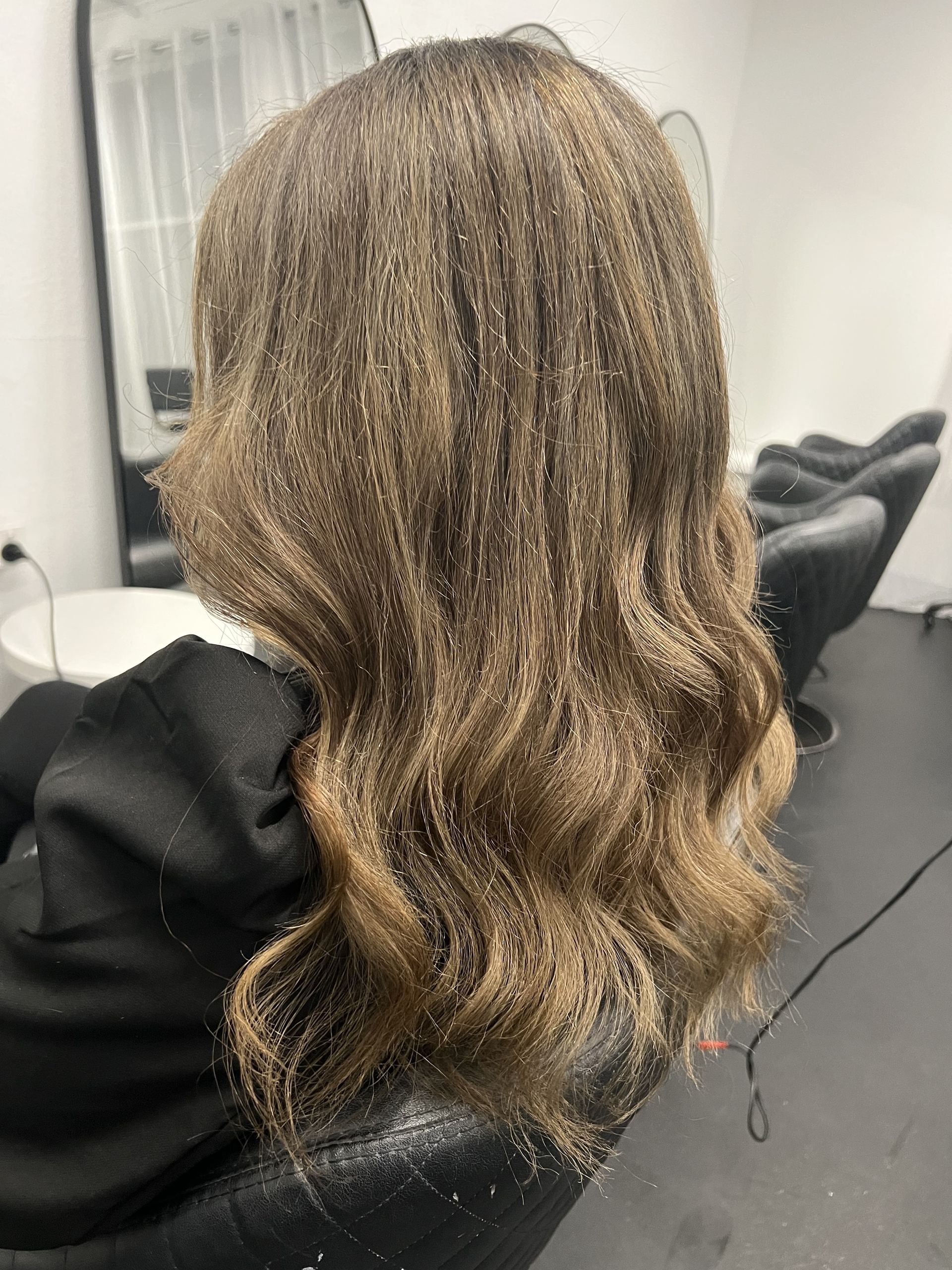 Wavy, light brown hair styled in loose curls at a salon chair, viewed from — Elysian Salon In Wyong, NSW behind.