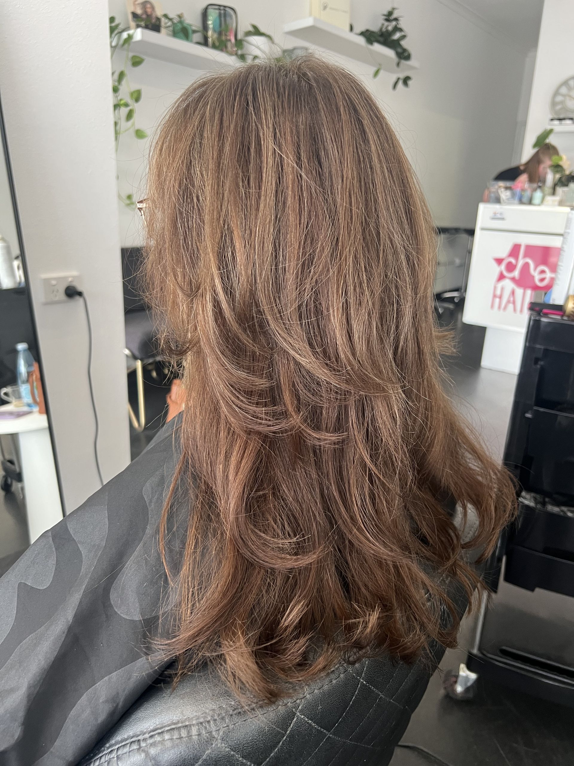 A Reflection of a Womans Hair in a Mirror — Choo Choo Hair and Beauty In Wyong, NSW Back view of shoulder-length layered brown hair in a salon chair, with a mirror and salon products nearby — Elysian Salon In Wyong, NSW
