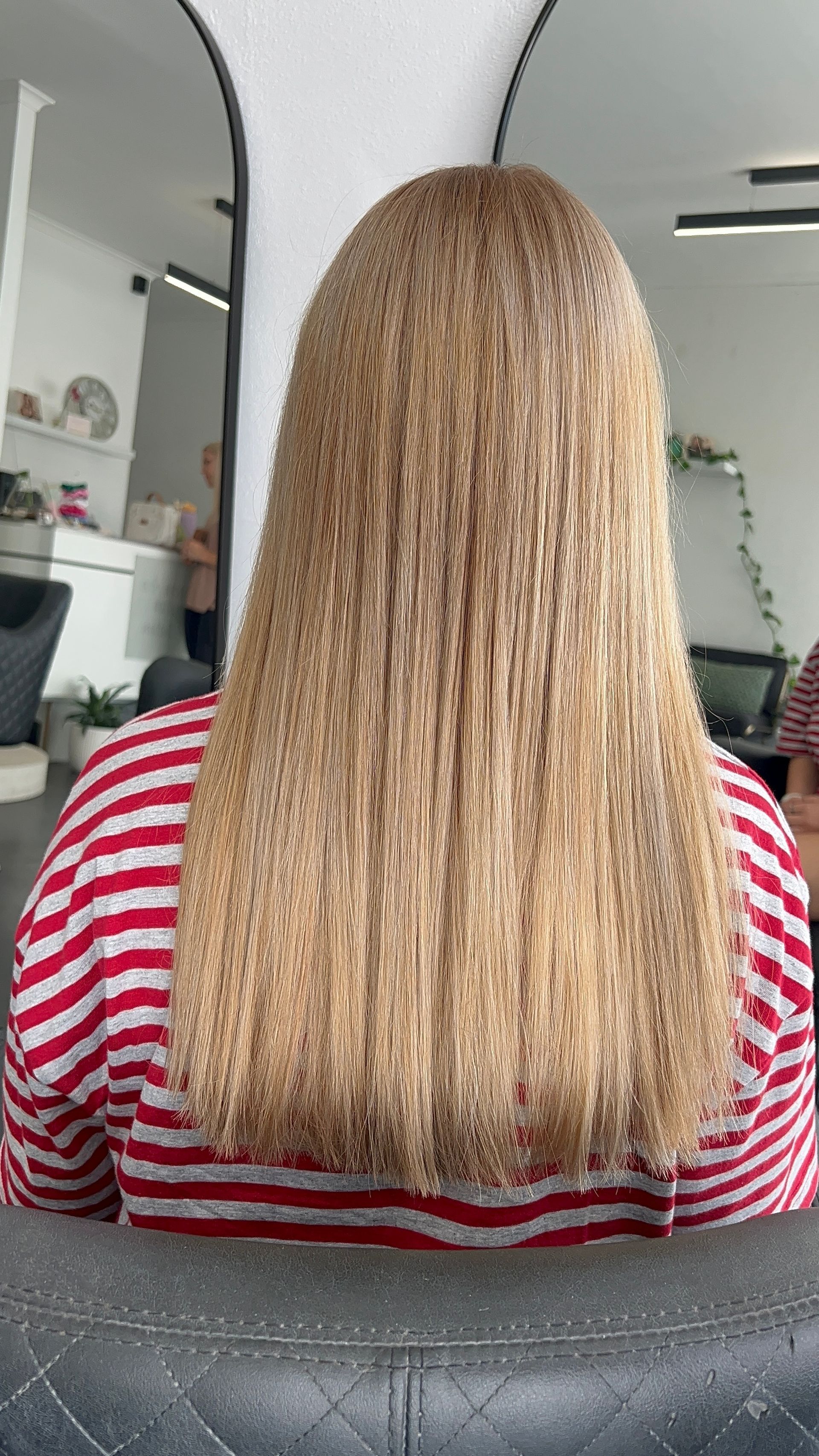 Back view of a person with long blonde curly hair in a red-and-white striped shirt, seated indoors — Elysian Salon In Wyong, NSW