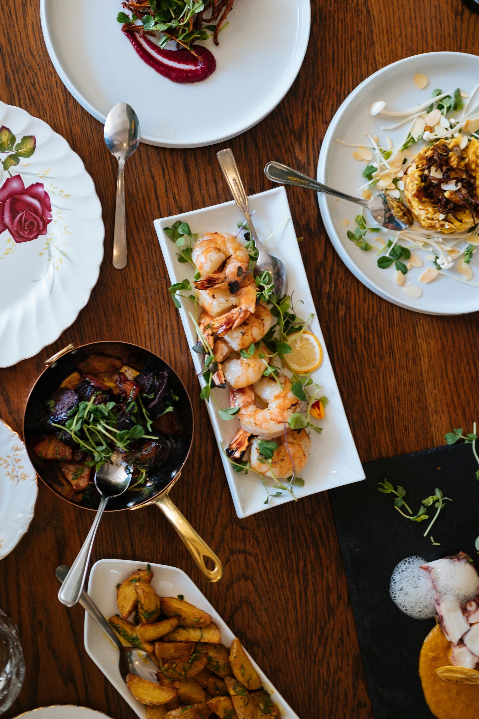 Overhead shot of a wooden table set with plates of gourmet food, including shrimp, potatoes, and pasta.