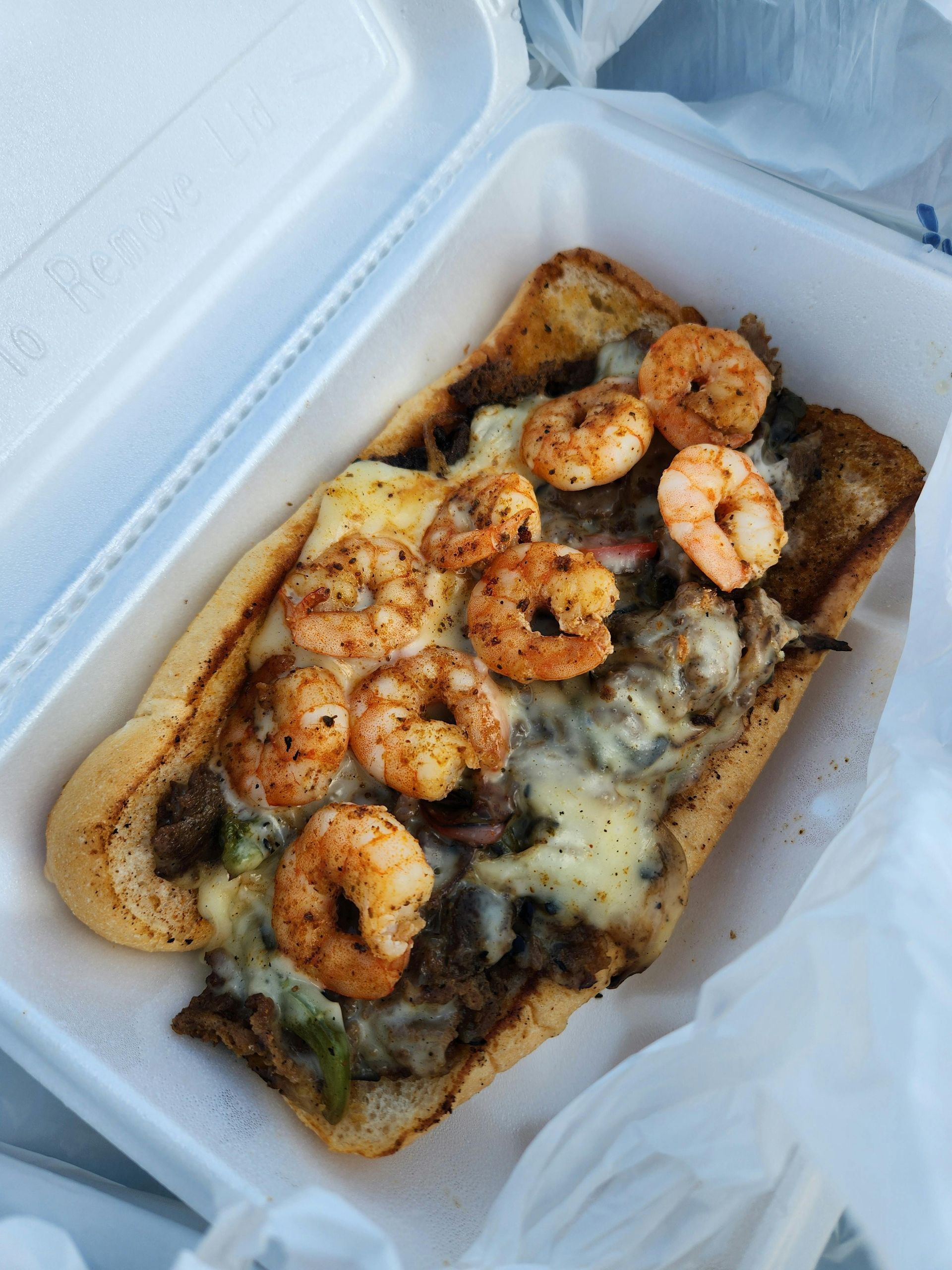 Sub sandwich with grilled shrimp, meat, and cheese in a styrofoam container.