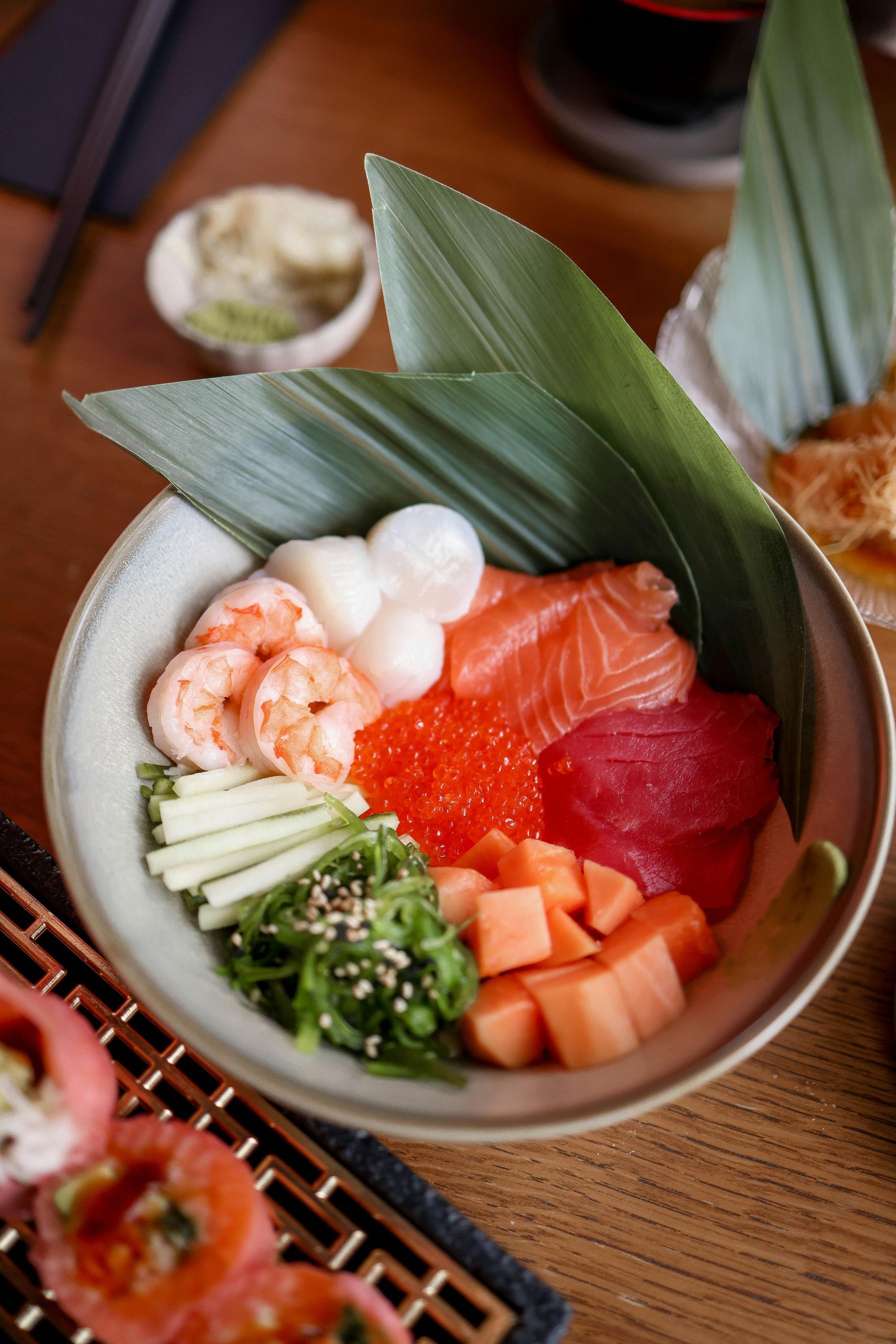 Sushi bowl with salmon, tuna, shrimp, roe, and other ingredients, garnished with a leaf.
