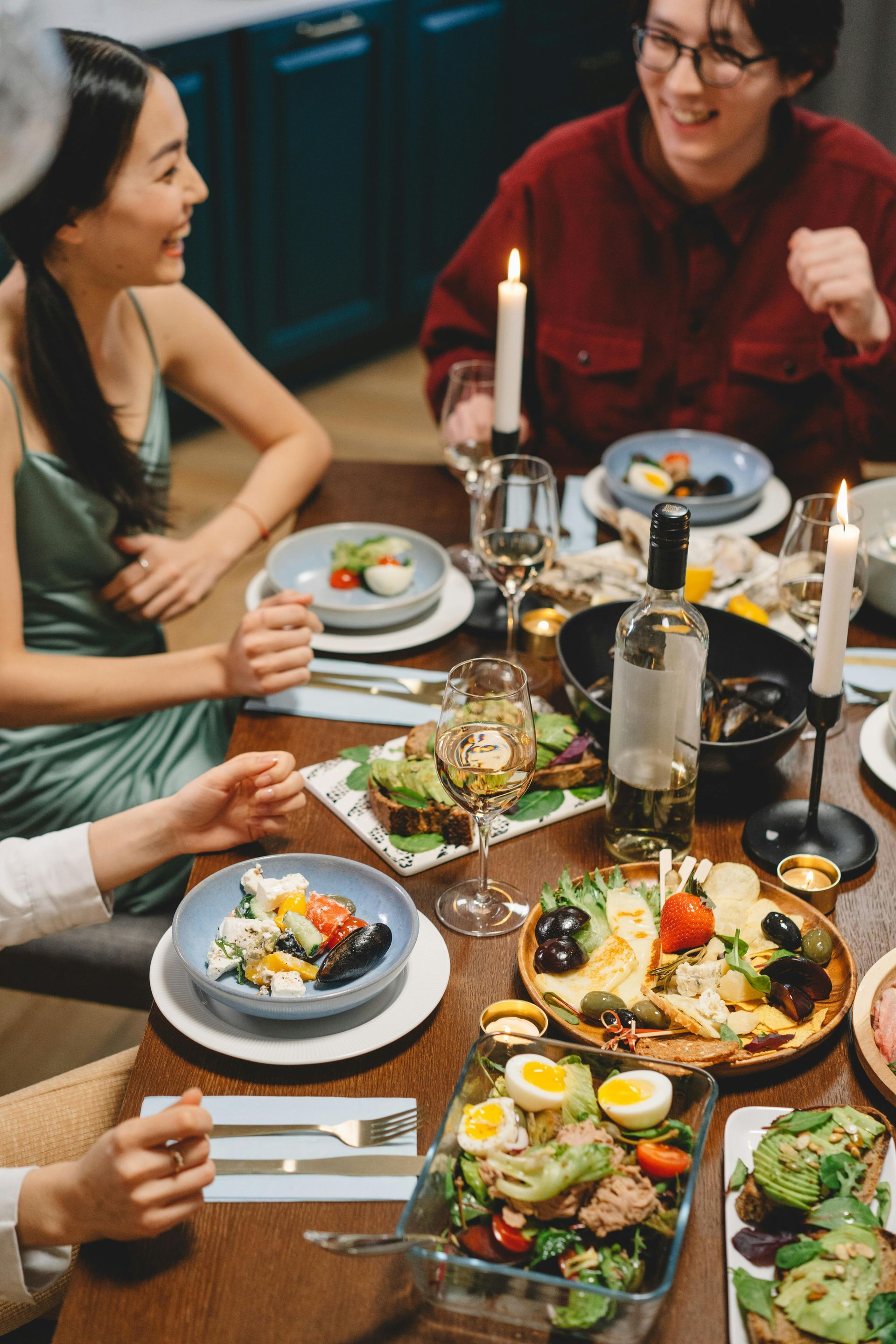 People gathered around a dining table laden with food, smiling and conversing. Candles illuminate the scene.