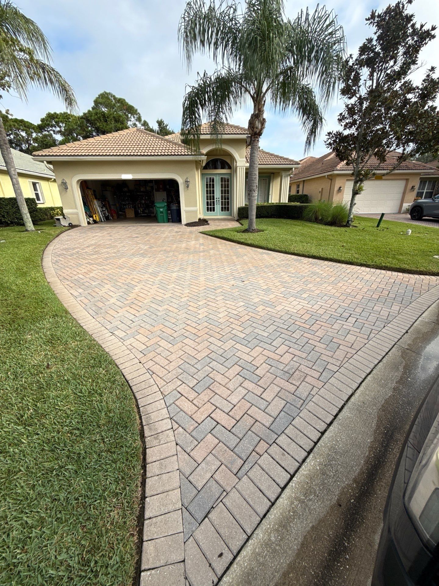 Brick driveway leading to a house, flanked by shrubs and trees under a sunny sky.
