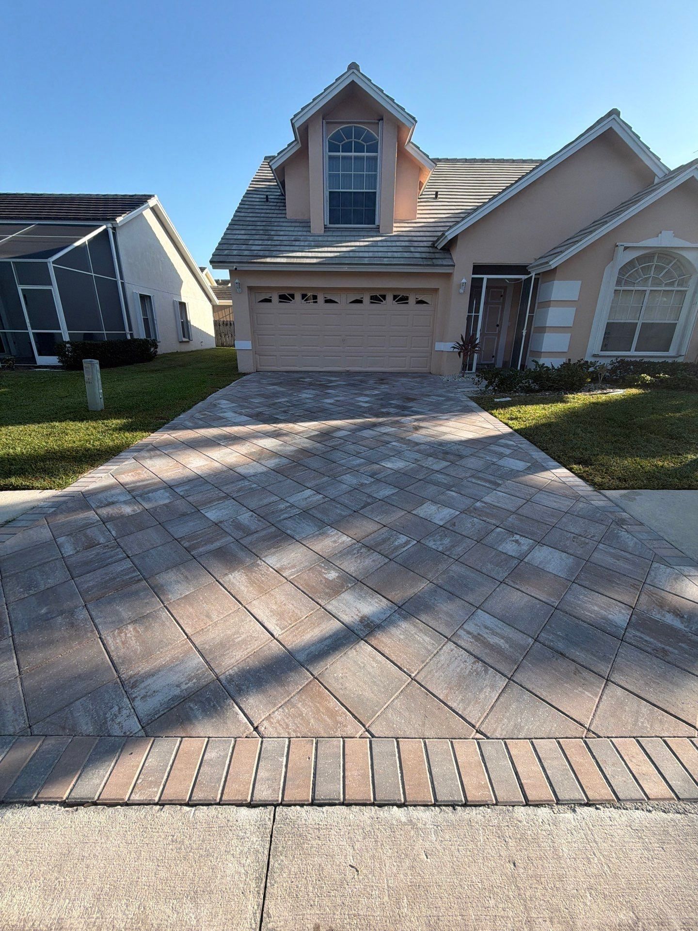 Paver driveway leading to a two-story beige house with a tan garage door and a dormer window.
