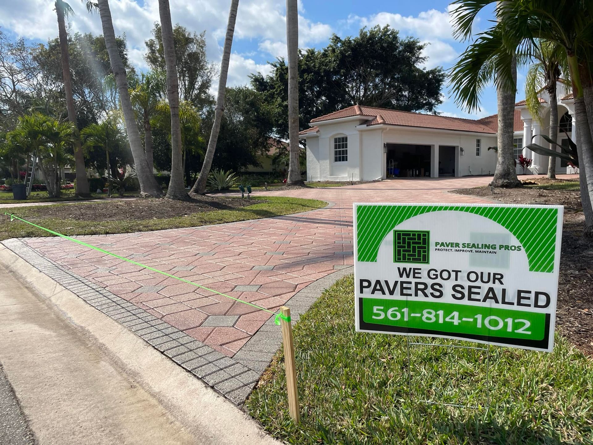 Paver sealing service sign in front of a brick driveway and house, with phone number in green.
