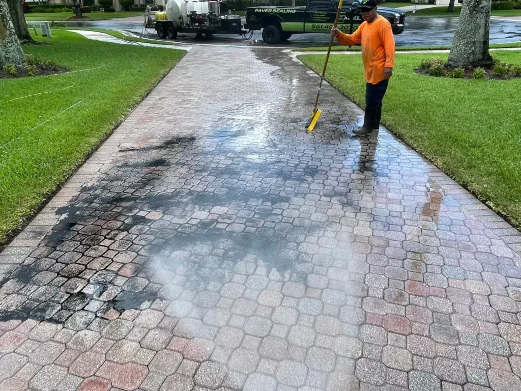 Man in orange scrubs sweeping a wet, patterned brick driveway; black truck and green lawn in background.