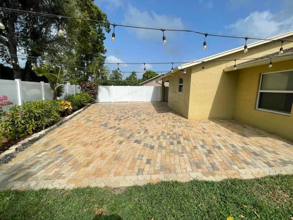 Backyard patio with brick pavers, yellow house, string lights, white fence, and greenery.