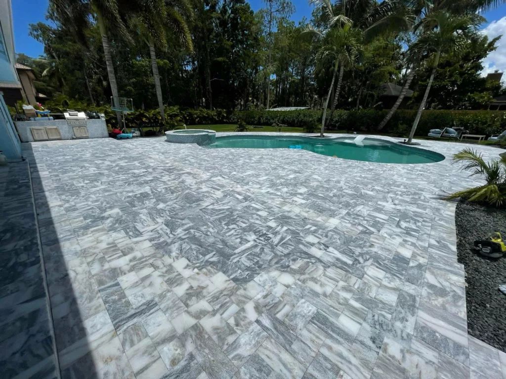 Stone-tiled patio with pool in background, palm trees, blue sky.
