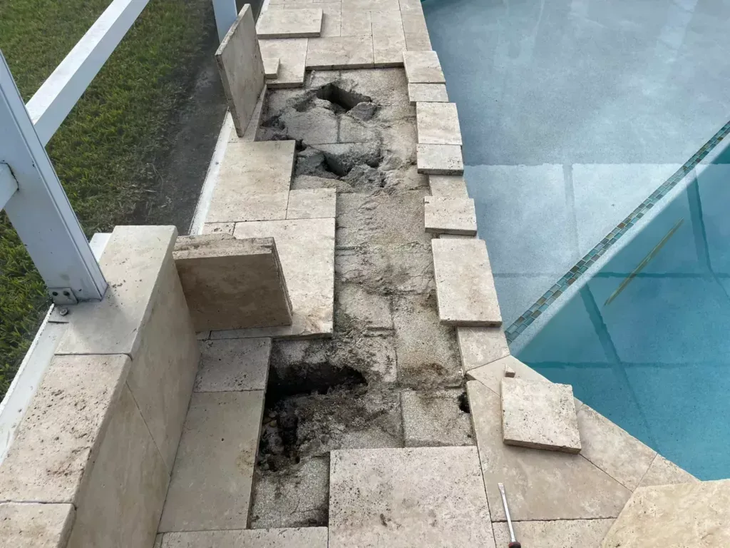 Damaged stone pool coping; missing sections exposing underlying structure, next to a pool and railing.