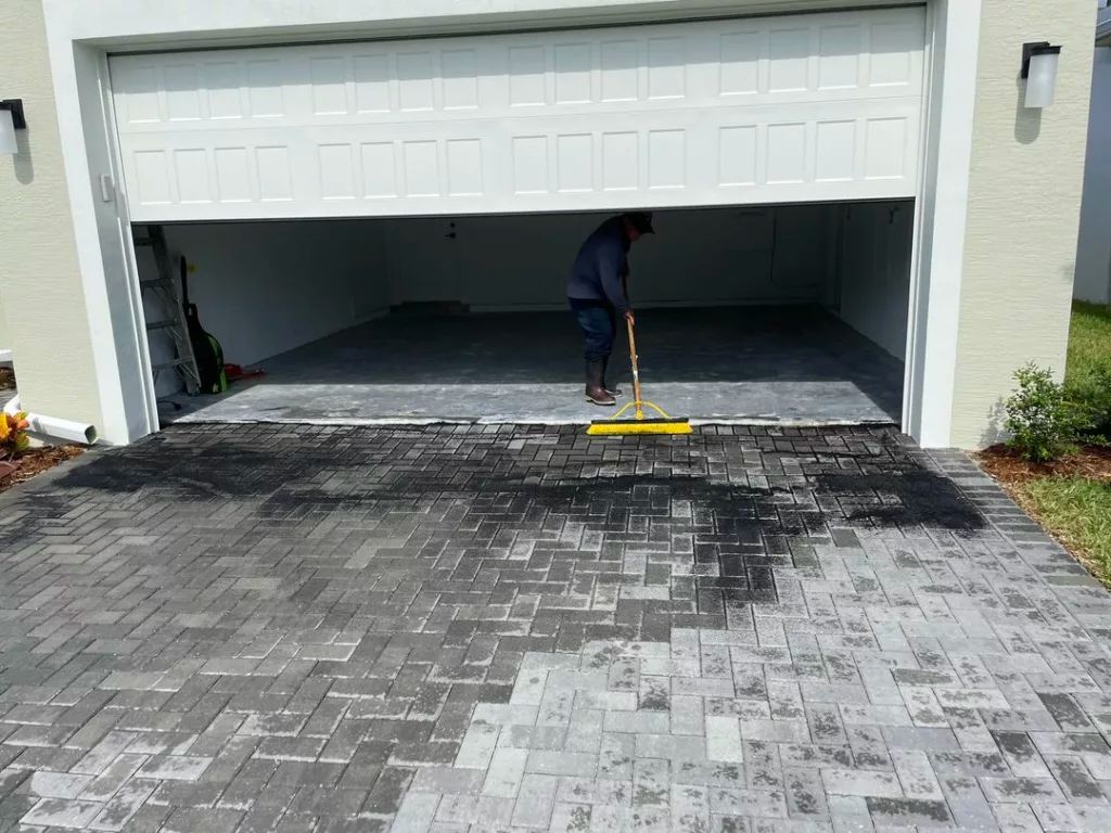 Person sweeping water on a brick driveway, in front of an open garage.
