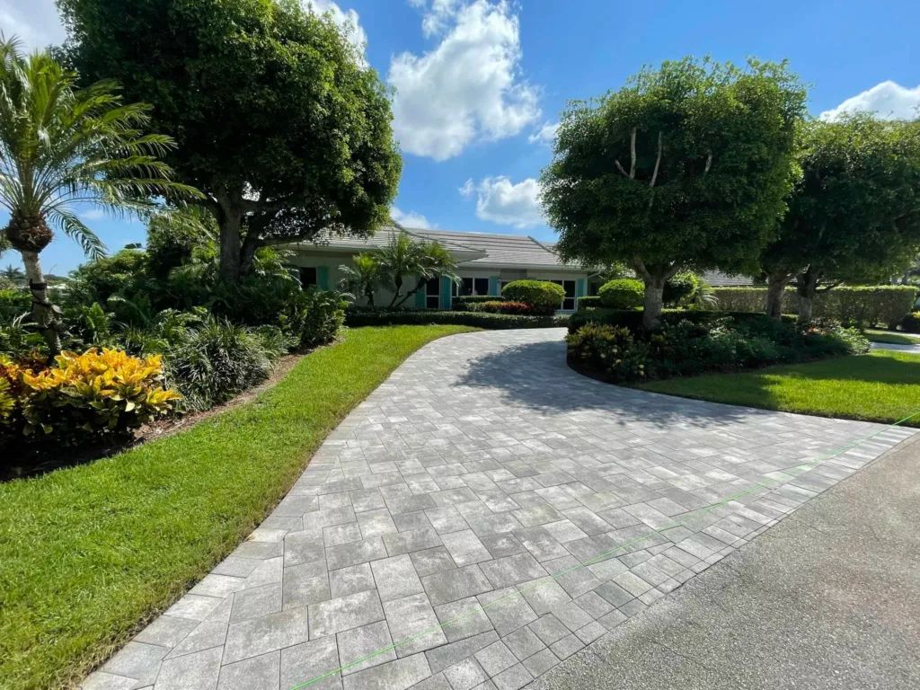 Driveway leading to a light-colored house with green landscaping under a blue sky with clouds.