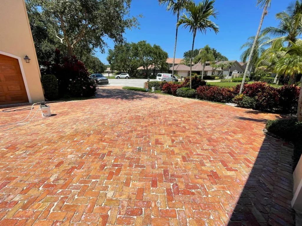 Brick driveway leading to a house, flanked by shrubs and trees under a sunny sky.