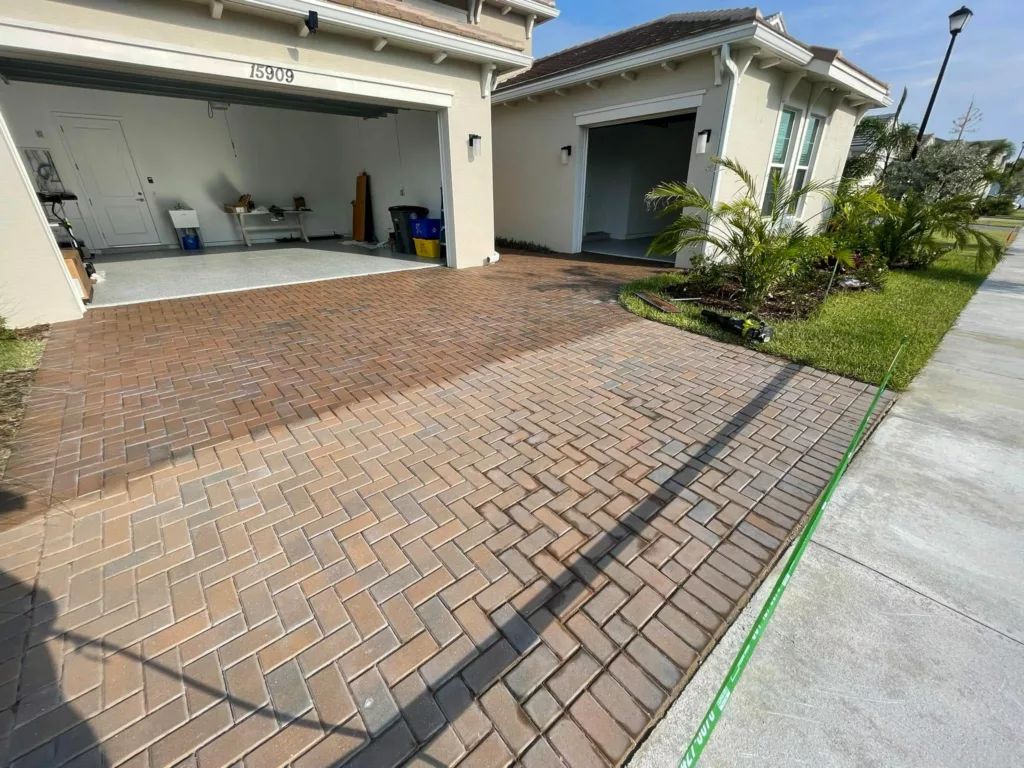 Brick paver driveway in front of a beige house with two garage doors and a small building with a window.