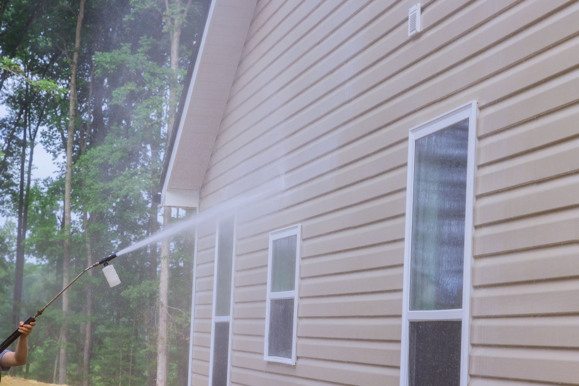 Person power washing the beige siding of a house with white trim.