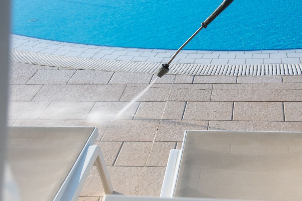 Pressure washing pool deck next to a blue pool, cleaning around white lounge chairs.