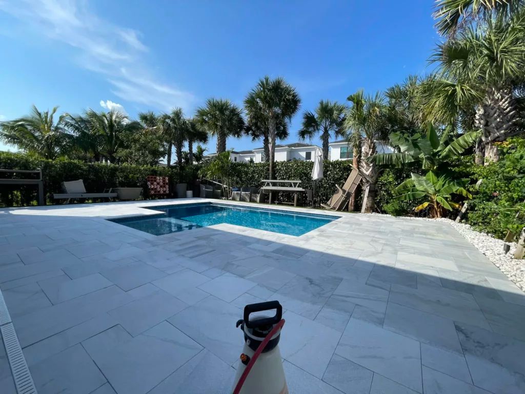 Pool with surrounding white stone patio, palm trees, and clear blue sky.