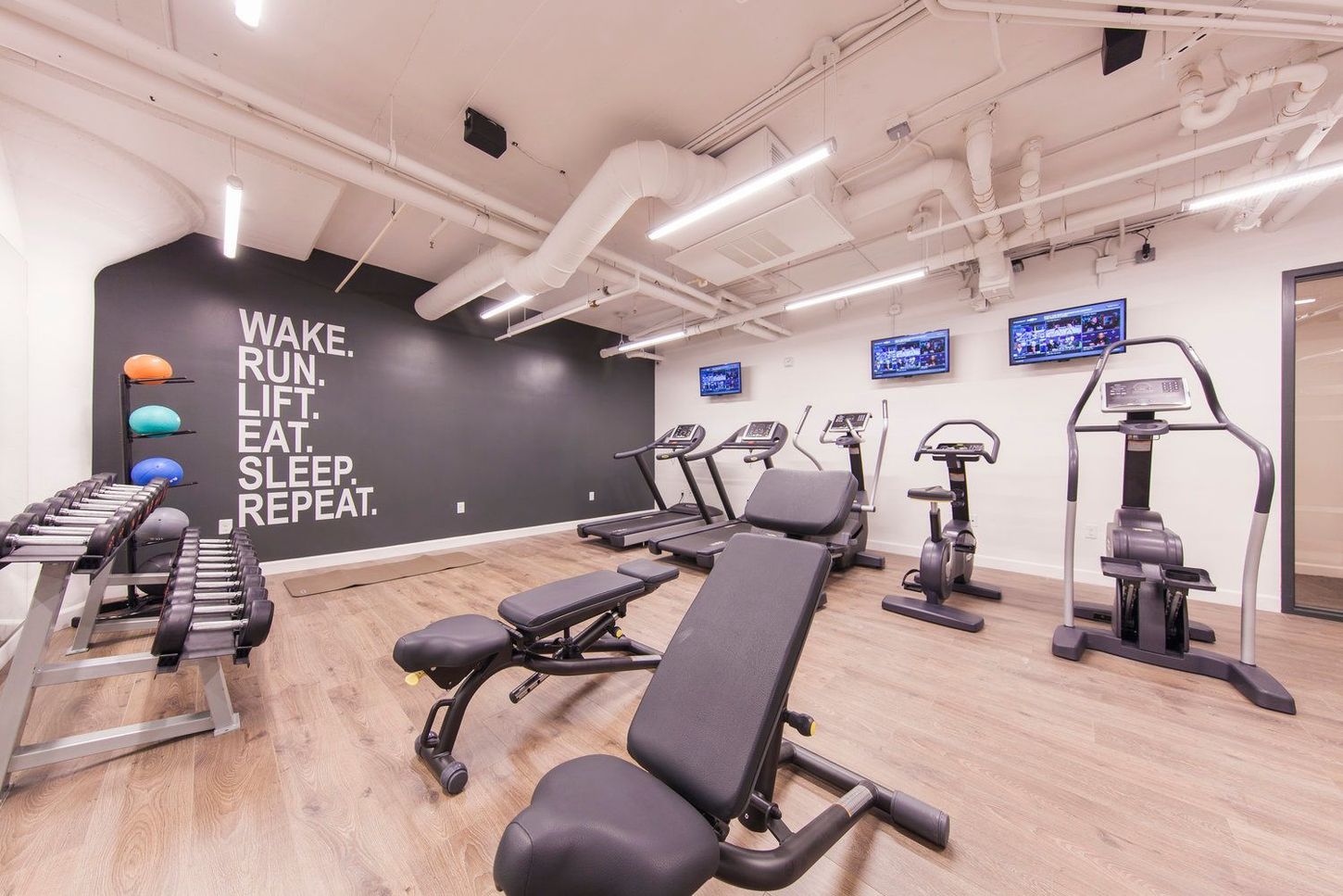 Gym with weights, benches, and exercise machines against a dark wall with motivational text; bright lighting.
