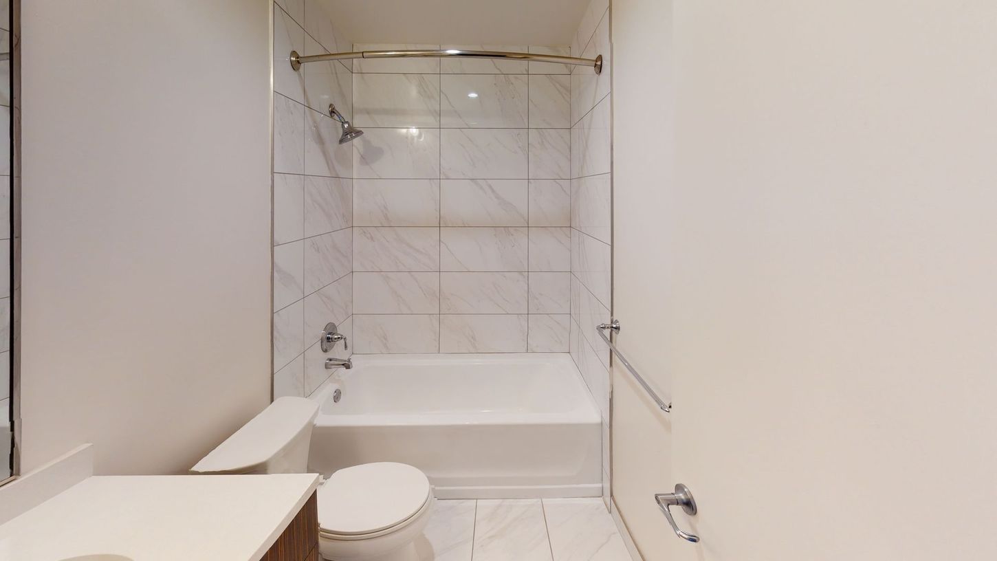 Bathroom with white tile, toilet, and bathtub. Silver fixtures and a grab bar are visible.
