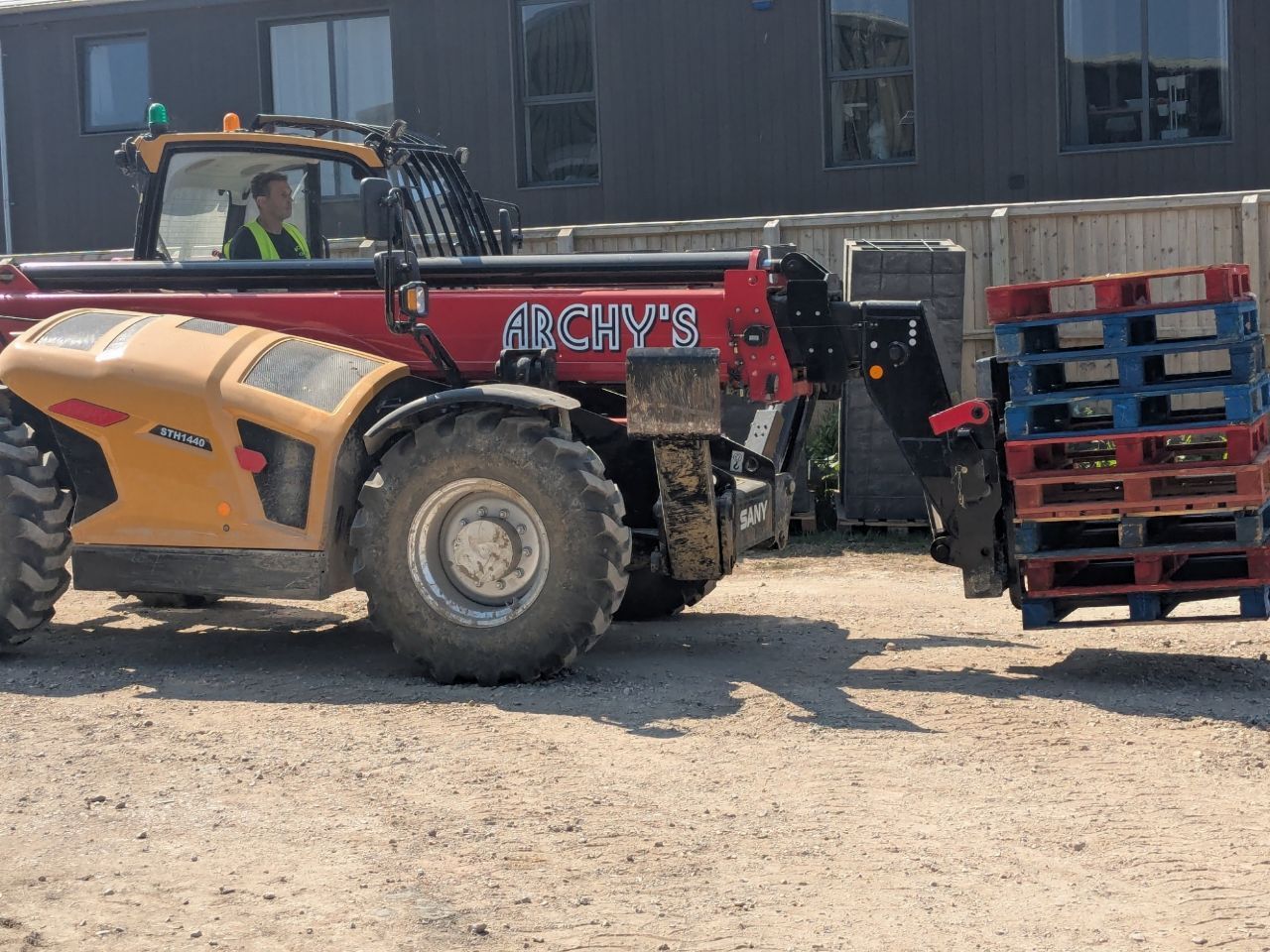 A red and yellow telehandler is carrying pallets in a dirt field.