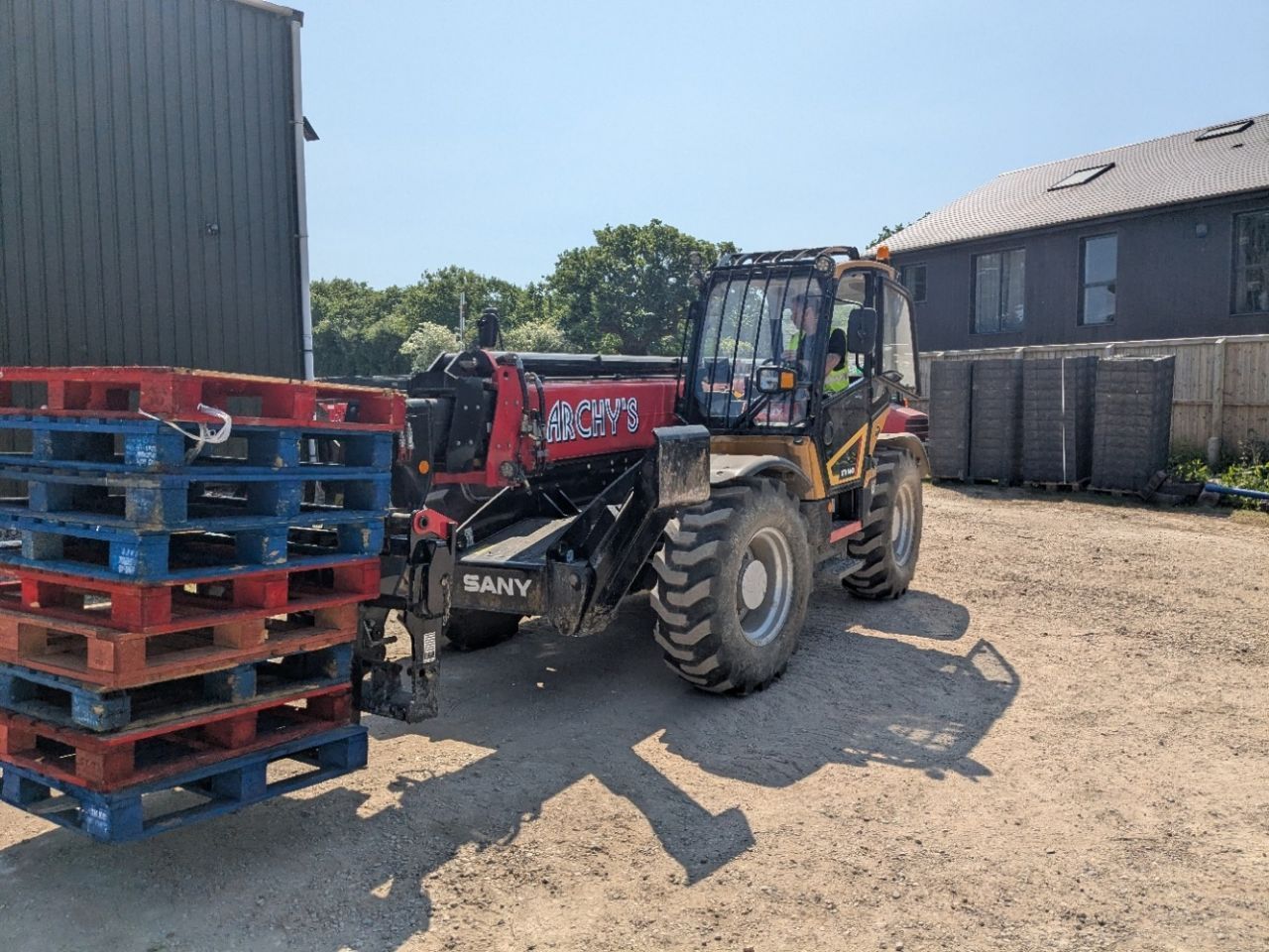 A red and yellow telehandler is carrying pallets in a dirt field.