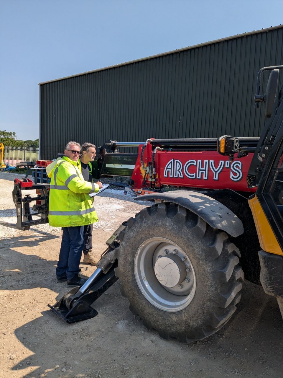 A man in a yellow jacket is standing next to a red telehandler that says archy 's on it.