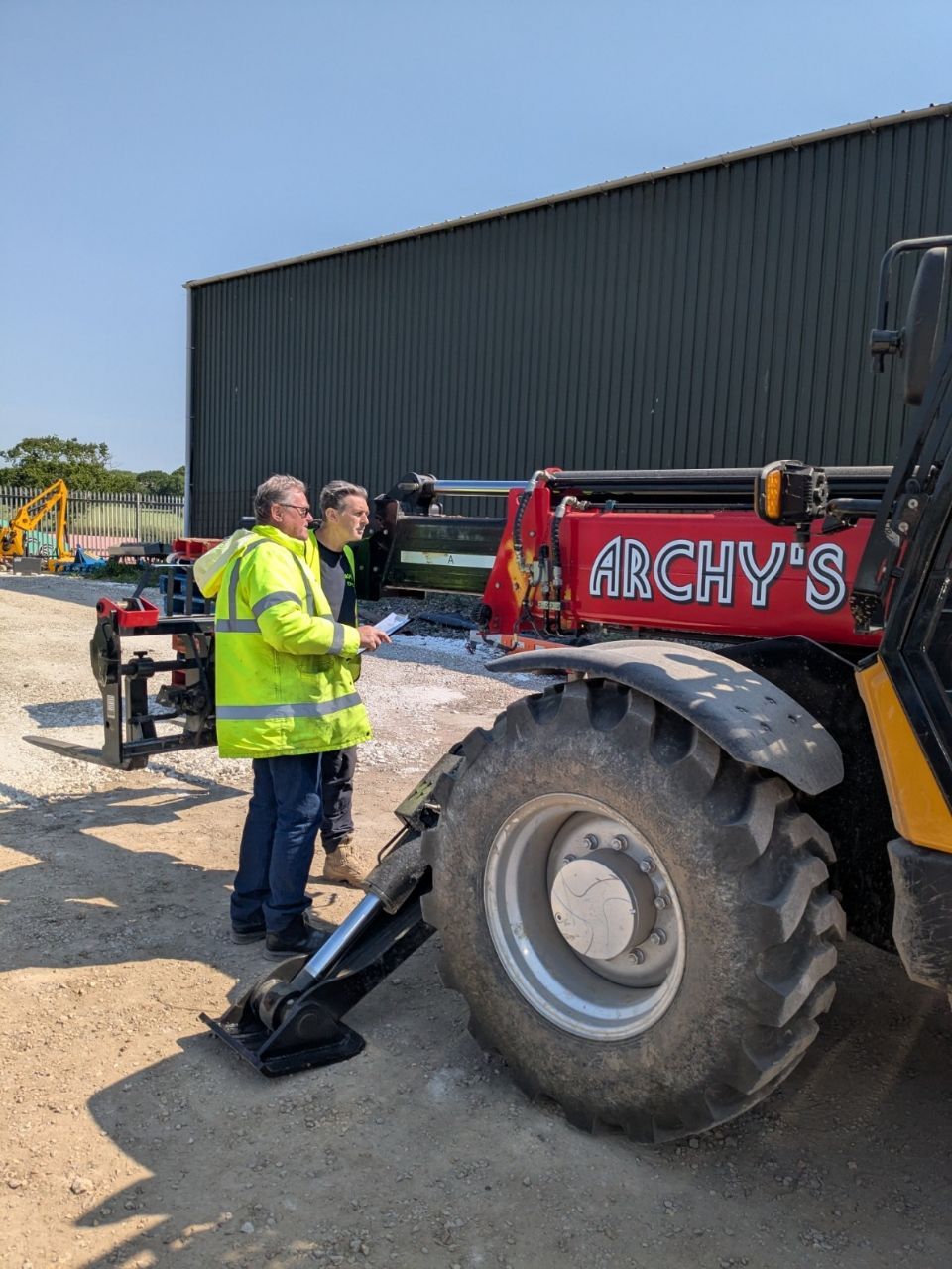 Two men are standing next to a red telehandler