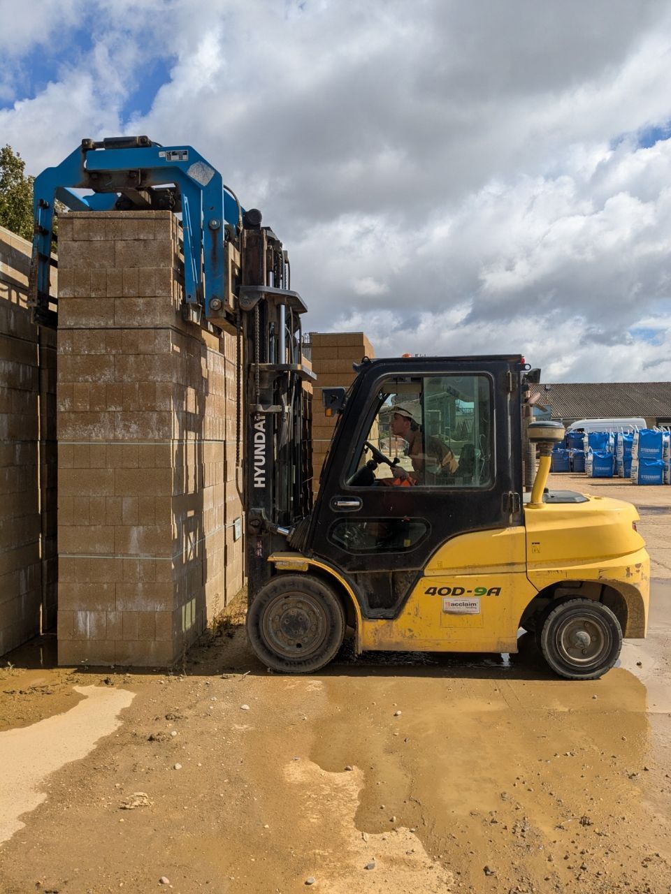 Yellow forklift with blue clamp lifting stacked concrete blocks; outdoor setting.